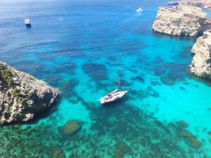 Private boat anchored in crystal-clear turquoise water with people snorkelling directly off the boat in Comino, Malta.