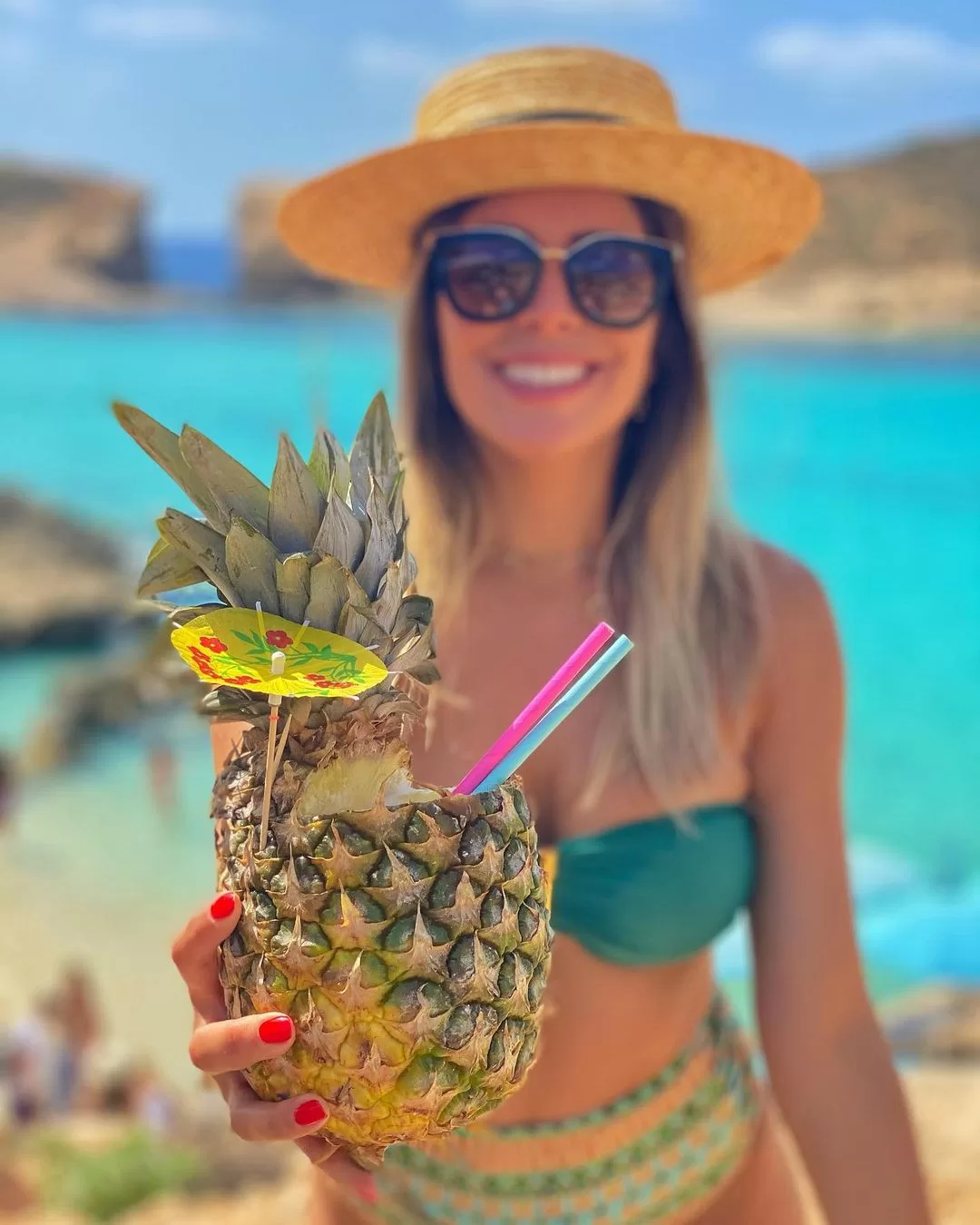 Woman holding a pineapple while relaxing in the turquoise waters of Comino, Malta near a boat.