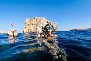 Person snorkeling in clear blue water in Comino, Malta, using a floating safety buoy while swimming away from a boat.