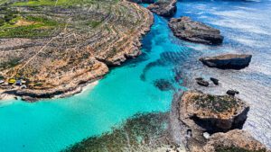 Aerial view of the Blue Lagoon at Comino Malta, showing turquoise waters, shallow areas, and boats surrounded by clear blue sea.