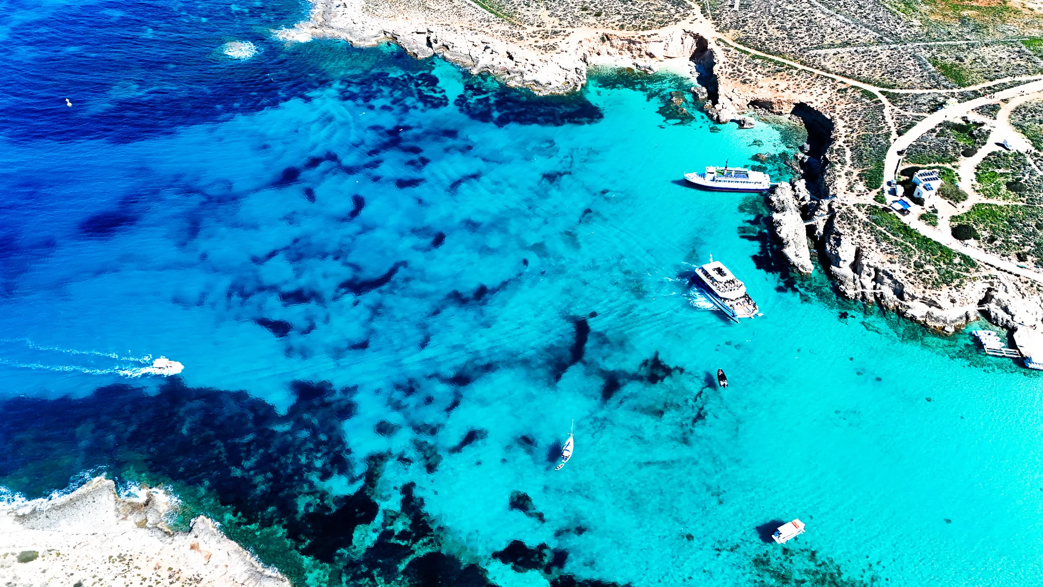 Aerial view of the Blue Lagoon at Comino Malta, showing vibrant turquoise waters, shallow areas, and boats surrounded by clear sea.