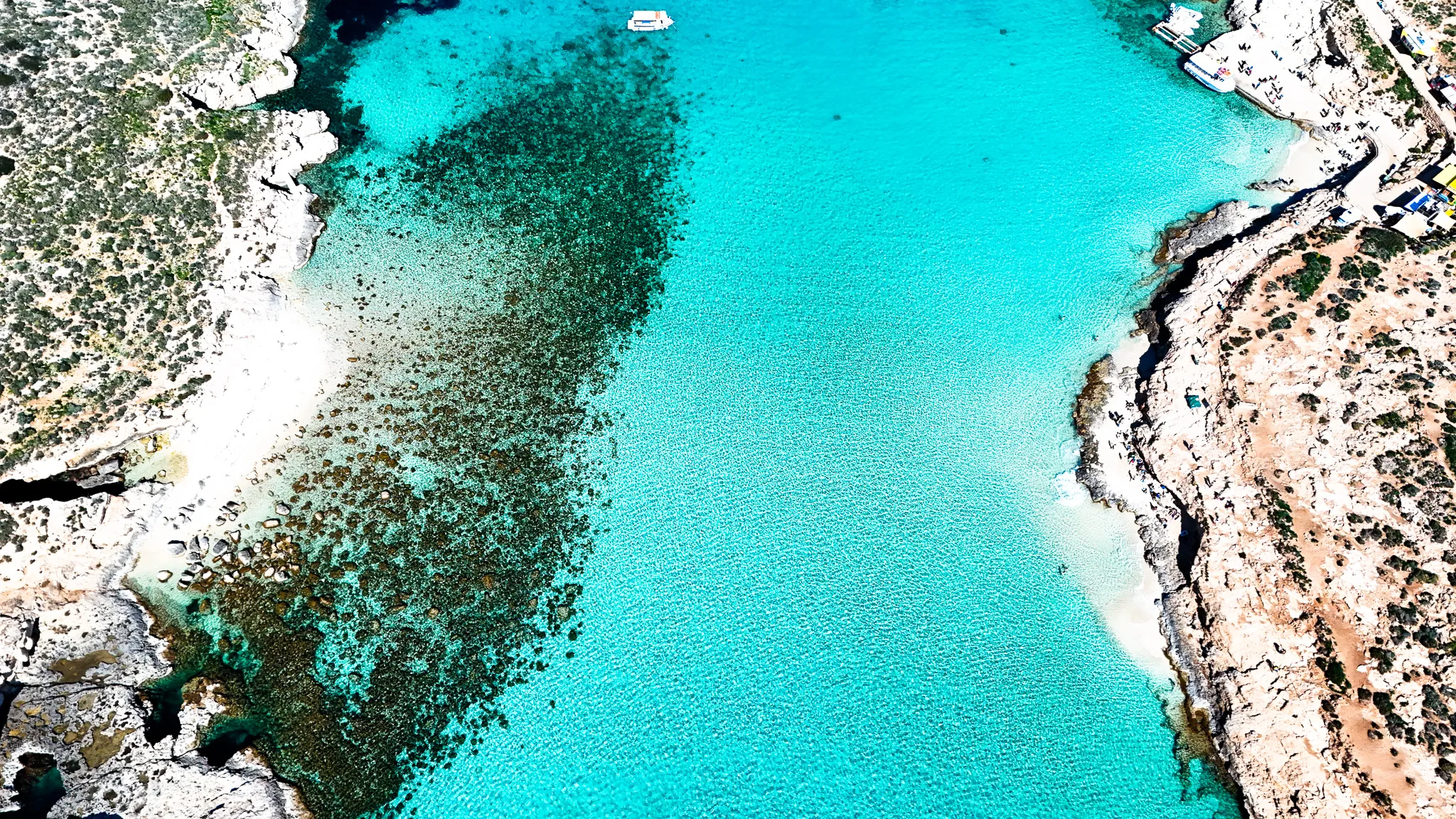 Drone view of the Blue Lagoon at Comino Malta, showing bright blue waters, shallow areas, and boats surrounded by clear sea.