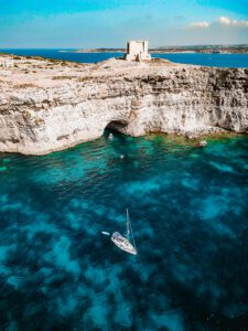 Drone shot of Crystal Lagoon in Comino Malta, showing deep blue water, cliffs, and boats surrounded by crystal clear sea.